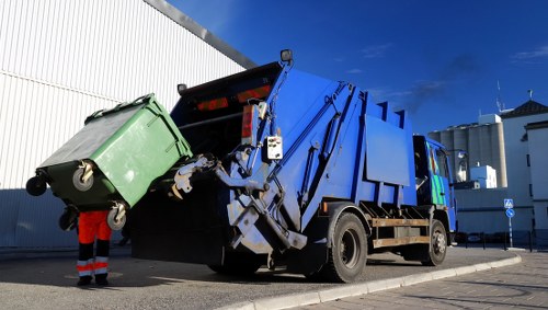 A blue garbage collection truck with a hydraulic lifting mechanism at the rear is actively engaged in emptying a green wheeled waste bin, which is positioned upside down as it is being emptied into the truck's hopper. The bin, made of durable plastic, has a textured surface and is secured with black rubber wheels. The scene takes place on a paved area next to a white industrial building, with part of the truck's cab visible on the right side, showing a glass window and side mirror. A worker, dressed in high-visibility orange trousers and a dark top, is standing nearby, assisting with the process. The background shows a bright blue sky with a few clouds, and the environment appears clean and organized, typical of an urban or commercial waste collection setting. This image illustrates a professional rubbish removal operation, representative of services offered by Pinner House Clearance in the local Pinner area, highlighting the efficient collection of household or commercial waste.