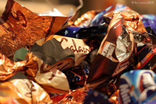 A close-up image showing a pile of crumpled foil wrappers and snack packaging materials, primarily in metallic copper, silver, and vibrant patterned designs. The wrappers are scattered and overlapping, with some partially unfolded, revealing glossy textures and bright colours. The background is blurred but suggests an indoor setting with warm ambient lighting. The clutter appears to be collected for rubbish removal purposes, hinting at a typical waste collection scene relevant to services like those offered by Pinner House Clearance in the local area. The focus is on the detailed surface textures of the wrappers, emphasizing their reflective properties and creased forms as part of general waste ready for disposal or collection.