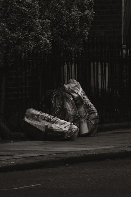 A black wheeled rubbish bin with a white label and the name 'St. John's' printed on the front, positioned on a paved sidewalk next to a curb in an outdoor urban setting at night. The bin is filled wit